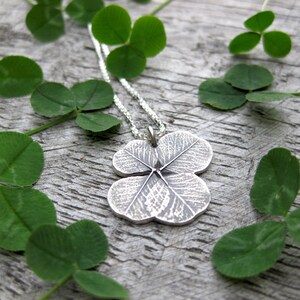 May include: A silver necklace with a four-leaf clover pendant. The pendant is detailed with veins and a textured surface. The necklace is displayed on a wooden surface with green clover leaves.