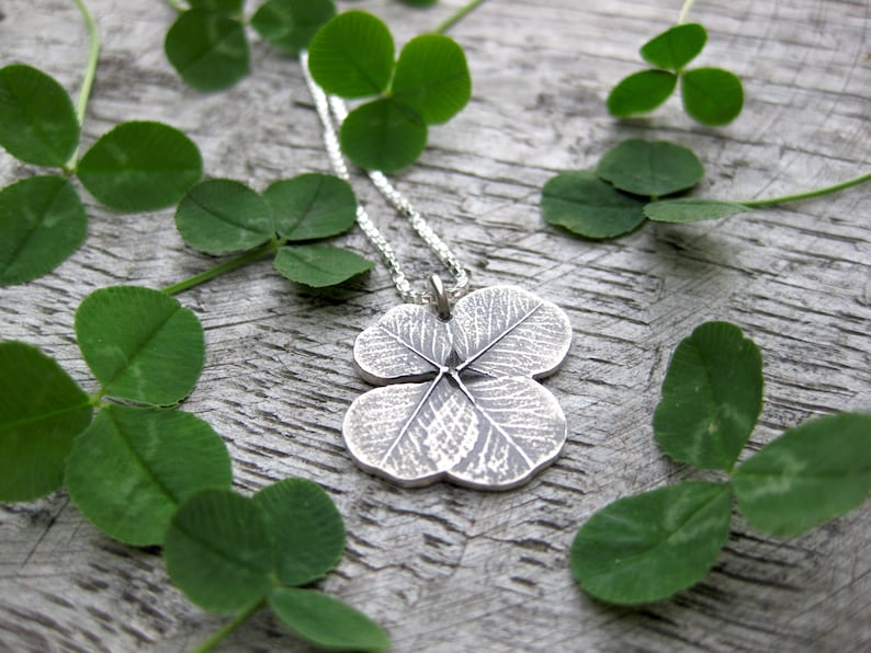 May include: A silver necklace with a four-leaf clover pendant. The pendant is detailed with veins and a textured surface. The necklace is displayed on a wooden surface with green clover leaves.