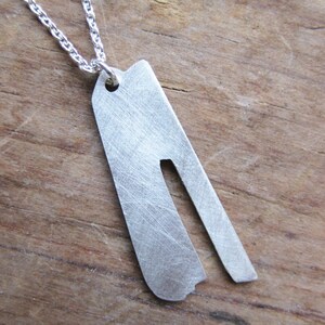 May include: A silver necklace with a pendant shaped like a long, thin rectangle with a cut-out in the middle. The pendant has a brushed finish.