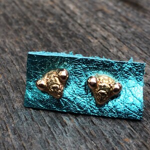 May include: A pair of gold-toned earrings with three small rose gold accents on each earring. The earrings are displayed on a turquoise leather background.