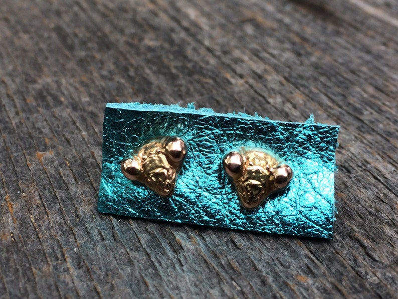 May include: A pair of gold-toned earrings with three small rose gold accents on each earring. The earrings are displayed on a turquoise leather background.