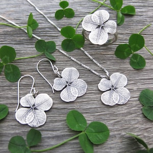 May include: A set of silver clover leaf jewelry, including a necklace, earrings, and a ring. The jewelry is displayed on a wooden surface with green clover leaves.