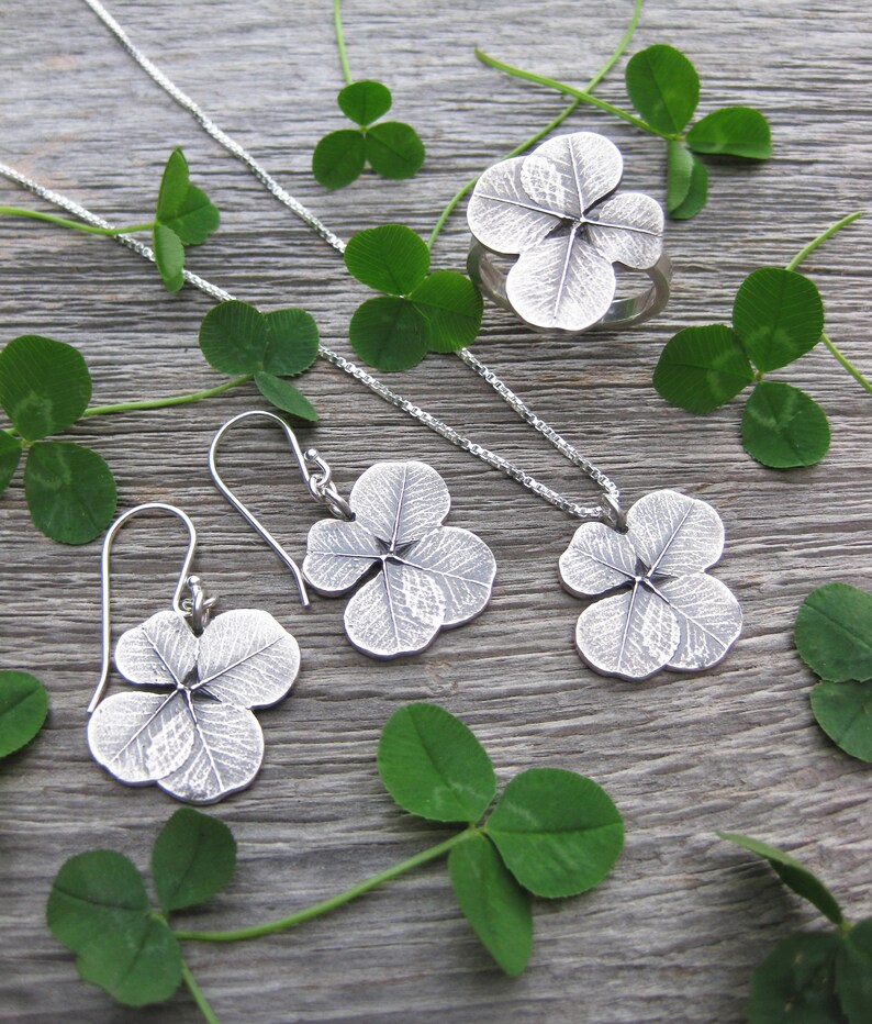 May include: A set of silver clover leaf jewelry, including a necklace, earrings, and a ring. The jewelry is displayed on a wooden surface with green clover leaves.