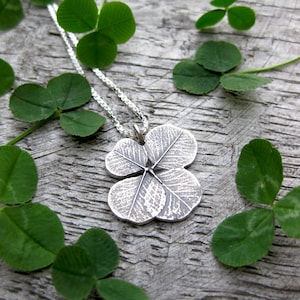 May include: A silver four-leaf clover pendant necklace with a detailed textured design, hanging on a silver chain against a background of green clover leaves.