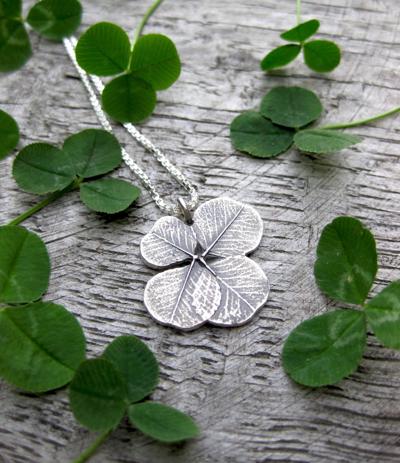 May include: A silver four-leaf clover pendant necklace with a detailed textured design, hanging on a silver chain against a background of green clover leaves.