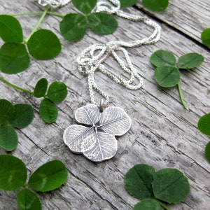 May include: A silver necklace with a four-leaf clover pendant. The pendant is detailed with the veins of the clover leaf. The necklace is surrounded by green clover leaves.