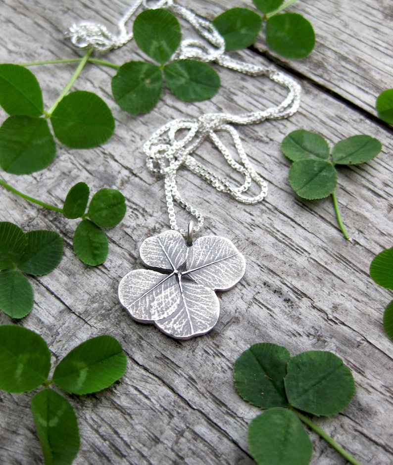 May include: A silver necklace with a four-leaf clover pendant. The pendant is detailed with the veins of the clover leaf. The necklace is surrounded by green clover leaves.
