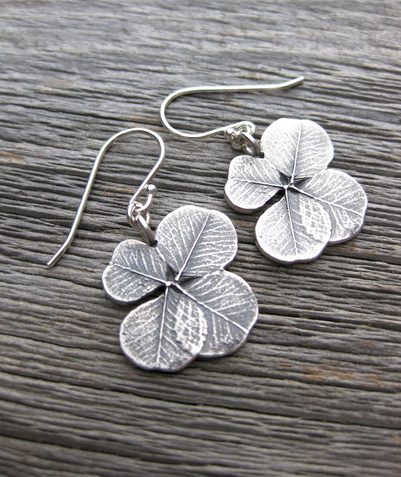 May include: A pair of silver earrings shaped like four-leaf clovers. The earrings have a textured, detailed design and are hanging from silver hooks.