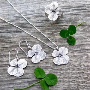 May include: A set of silver clover leaf jewelry, including a necklace, earrings, and a ring. The jewelry is displayed on a wooden surface with real clover leaves.