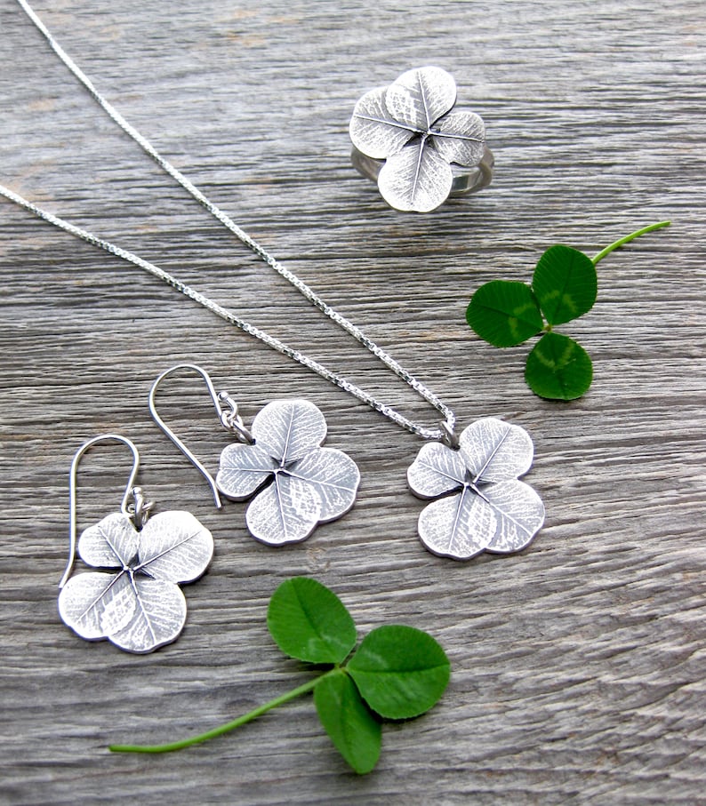 May include: A set of silver clover leaf jewelry, including a necklace, earrings, and a ring. The jewelry is displayed on a wooden surface with real clover leaves.