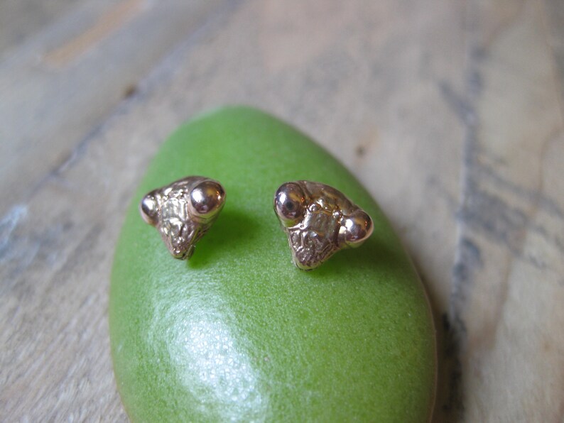 May include: A pair of gold-toned earrings shaped like the head of a praying mantis. The earrings are small and delicate, with detailed features.