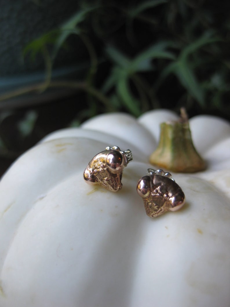 May include: A pair of gold-toned stud earrings with a textured, organic shape. The earrings are set against a white background.