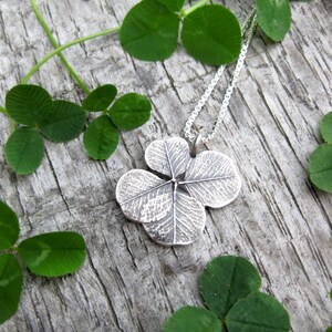 May include: A silver four-leaf clover pendant necklace on a silver chain, resting on a wooden surface with green clover leaves.