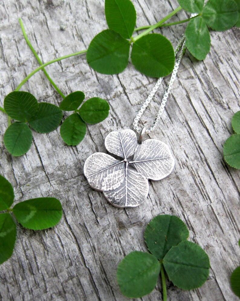 May include: A silver four-leaf clover pendant necklace on a silver chain, resting on a wooden surface with green clover leaves.