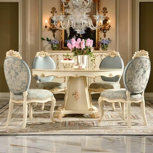 May include: A classic dining set with a round table and four chairs. The table and chairs are cream with gold detailing. The chairs have patterned backs and seats. A chandelier hangs above the table, and a floral arrangement is in the centre.