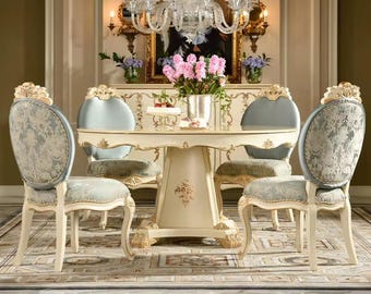 May include: A formal dining set featuring a round table and four chairs. The table and chairs are cream-colored with gold accents. The chairs have patterned backs and seats. A chandelier hangs above the table, and a floral arrangement sits in the center.