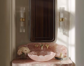 May include: A luxurious bathroom vanity with a pink marble countertop and a matching scalloped sink. The vanity has gold-colored metal legs and a large rectangular mirror. Two glass sconces with gold accents flank the mirror. A vase of roses and a candle sit on the countertop.