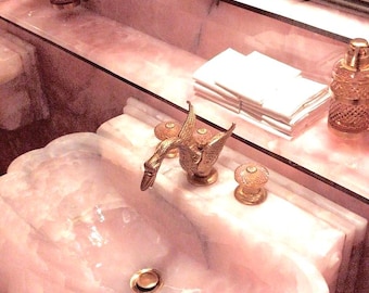 May include: A luxurious pink marble bathroom sink with a gold swan-shaped faucet. The sink is surrounded by a matching countertop, mirrors, and decorative bottles. A stack of white towels sits on the counter.