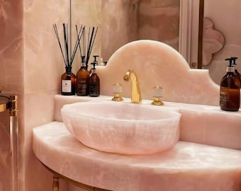 May include: A pink onyx bathroom vanity with a white porcelain sink and gold faucet. The vanity has a curved countertop and gold legs. There are three bottles of liquid soap on the countertop.