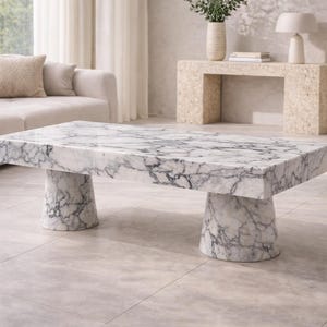 Puede incluir: Una mesa de centro rectangular de mármol con un patrón veteado gris y blanco. La mesa tiene una tapa gruesa y dos bases cilíndricas. La mesa está en un salón moderno.