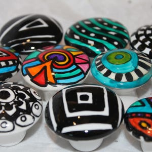 May include: A collection of ten ceramic drawer knobs with various abstract designs. The knobs are painted in bright colors, including black, white, red, blue, green, yellow, and purple. The designs are geometric and whimsical.