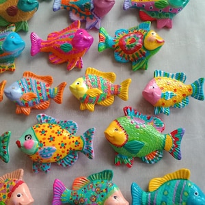 May include: A collection of colorful, hand-painted ceramic fish figurines. Each fish features unique patterns and vibrant colors, including pink, yellow, blue, and green. The fish have smiling faces and are arranged in a scattered pattern.