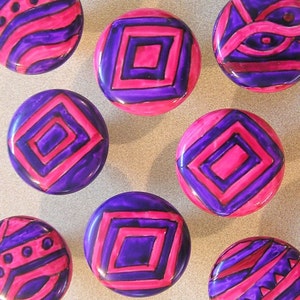 May include: Nine round, purple and pink painted ceramic knobs with geometric designs. The knobs have a glossy finish.