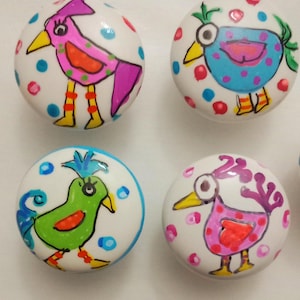 May include: Four white ceramic cabinet knobs with hand-painted bird designs. Each knob features a different colorful bird illustration with polka dots. The birds are in various colors, including pink, blue, green, and purple.