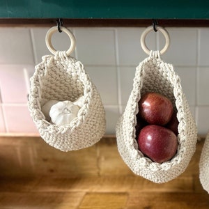 Könnte beinhalten: Drei hängende gehäkelte Körbe mit verschiedenen Produkten darin. Der Korb links enthält eine Knoblauchzehe, der mittlere Korb enthält rote Äpfel und der rechte Korb enthält Kartoffeln.