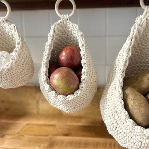 May include: Three white crocheted hanging baskets with different produce inside. The left basket holds a single garlic bulb, the middle basket holds three red apples, and the right basket holds five brown potatoes.