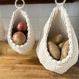 May include: Three white crocheted hanging baskets with different produce inside. The basket on the left contains a single garlic bulb. The middle basket contains three red apples. The basket on the right contains five brown potatoes.