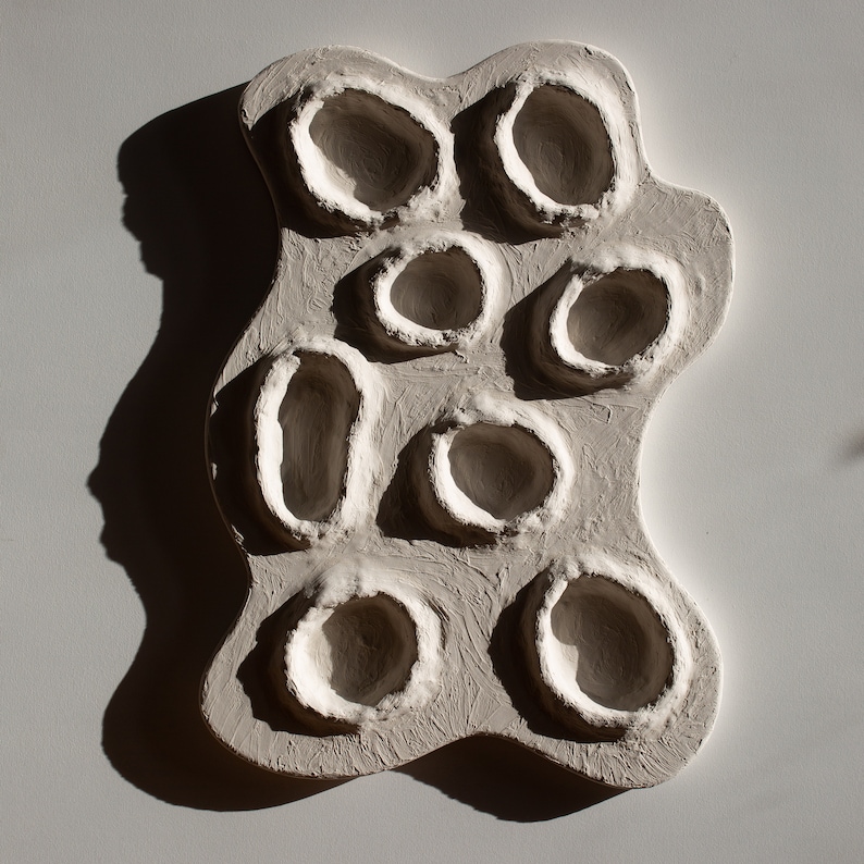 Pu&ograve; includere: Una scultura da parete in ceramica bianca con una superficie testurizzata e pi&ugrave; incavi circolari.