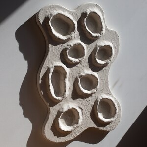 Pu&ograve; includere: Arte da parete in ceramica bianca con una superficie strutturata e pi&ugrave; rientranze circolari.