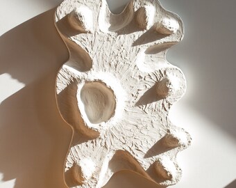 Può includere: Scultura da parete in ceramica bianca con una forma astratta e organica. La scultura ha una superficie testurizzata e diversi rilievi e rientranze.