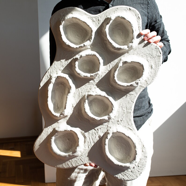 Pu&ograve; includere: Una scultura da parete bianca di forma organica con otto rientranze circolari. La scultura &egrave; realizzata in un materiale strutturato e ha un aspetto grezzo e incompiuto.