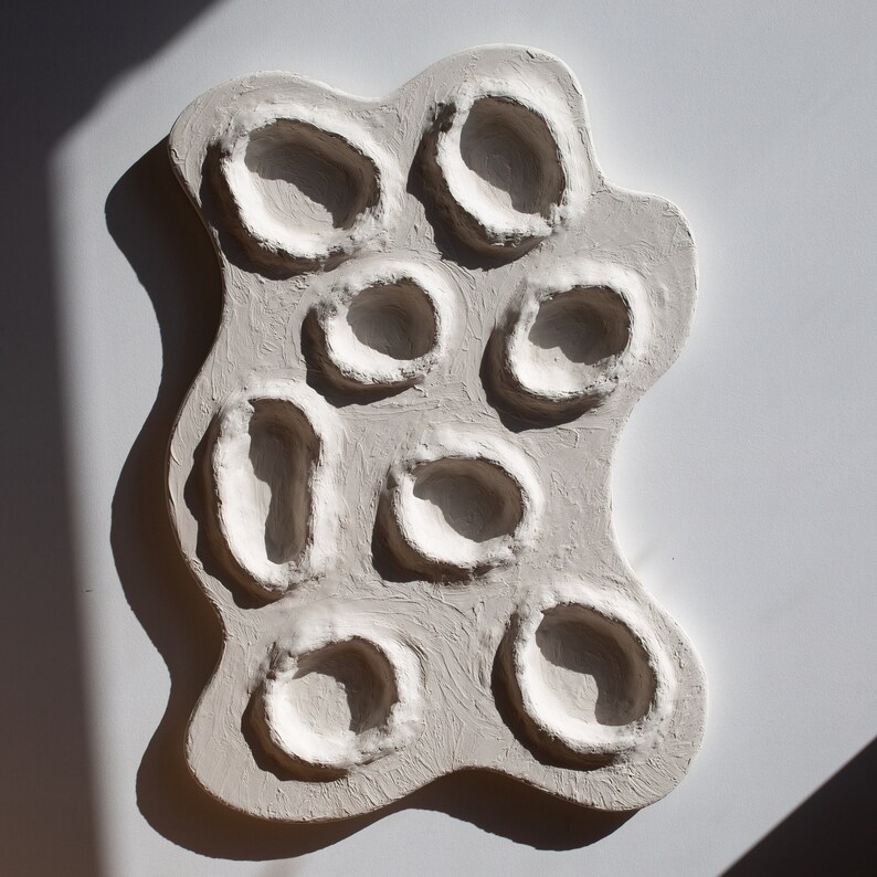 Pu&ograve; includere: Arte da parete in ceramica bianca con una superficie strutturata e pi&ugrave; incavi circolari.