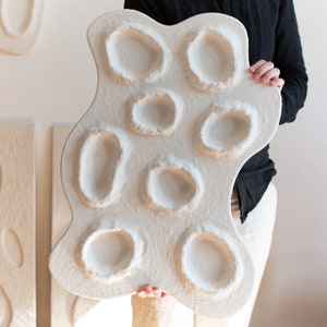 Pu&ograve; includere: Una scultura da parete bianca di forma organica con pi&ugrave; rientranze circolari. La scultura &egrave; realizzata in un materiale strutturato e ha un aspetto grezzo e fatto a mano.