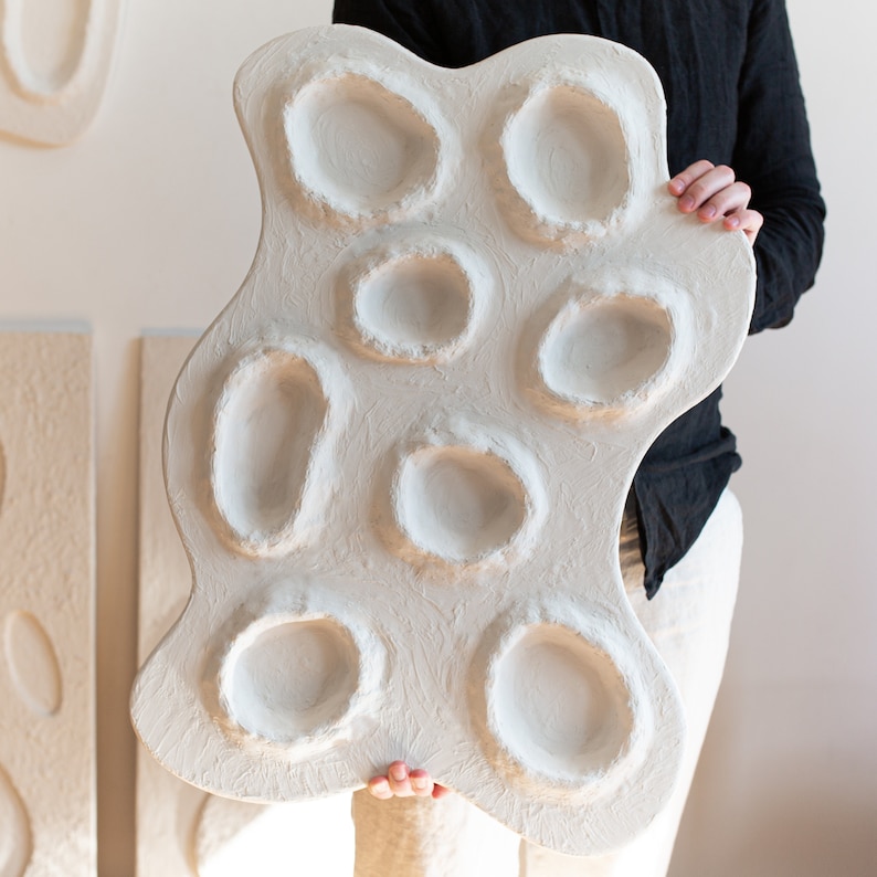 Pu&ograve; includere: Una scultura da parete bianca di forma organica con pi&ugrave; rientranze circolari. La scultura &egrave; realizzata in un materiale strutturato e ha un aspetto grezzo e fatto a mano.