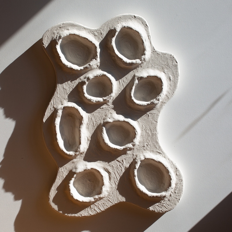 Pu&ograve; includere: Scultura da parete in ceramica bianca con una forma organica unica e multiple rientranze circolari.