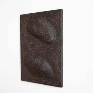 Pu&ograve; includere: Una scultura da parete in legno marrone scuro con due forme ovali in rilievo. La superficie della scultura ha un aspetto testurizzato e scolpito.