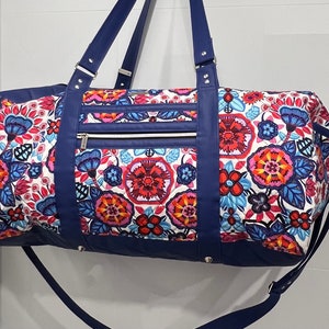 Op de afbeelding: Een blauwe reistas met een witte bloemenprint, donkerblauwe leren afwerking en een ritssluiting. De tas heeft een handvat en een schouderband.
