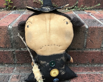 May include: A stuffed fabric doll shaped like a witch, featuring a tan face with stitched eyes and mouth, wearing a black hat and black dress adorned with three round buttons in green, yellow, and blue. The doll holds a small twig broom with straw.