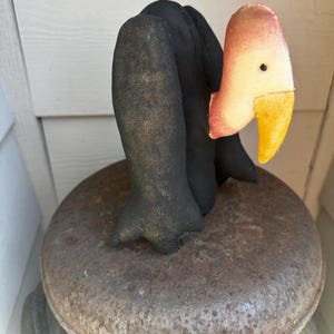 May include: A stuffed fabric vulture figurine featuring a black body with textured fabric wings, a pale pinkish-tan neck, and a bright yellow beak. The bird has a small black bead eye and is positioned in an upright, standing posture.