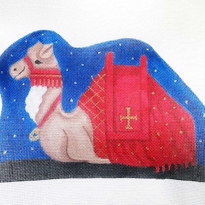 May include: A camel with a red and gold saddle and a blue blanket with white stars. The camel is kneeling and looking to the left.