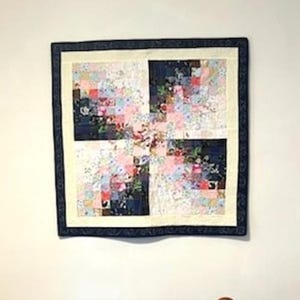 May include: A square wall quilt featuring a patchwork design of small fabric squares in shades of pink, white, and dark navy blue. The quilt has a cream-colored inner border and a dark navy blue outer border with a subtle repeating pattern.