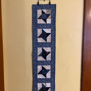 May include: A blue quilted wall hanging with five star-shaped designs. Each star is a mix of dark blue and patterned fabrics, set against a white background. The hanging is suspended from a decorative metal bracket.