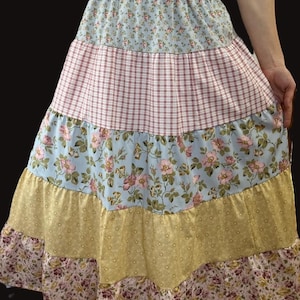 May include: A tiered skirt with a patchwork design. The skirt features layers of fabric in various patterns, including floral prints, gingham, and solid colors. The color palette includes light blue, pink, yellow, and burgundy. The skirt has an elastic waistband.