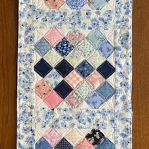 May include: A quilted table runner featuring a patchwork design with squares of pink, blue, and navy fabrics. The runner has a floral patterned border in shades of blue and white, adding a touch of elegance. The runner measures approximately 12 inches wide and 30 inches long.