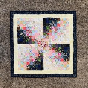 May include: A handmade patchwork quilt with a floral pattern. The quilt features a central design of squares in various colors, including pink, blue, and green. It has a cream-colored border and a dark blue outer border with a decorative swirl pattern.