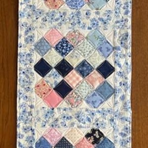 May include: A quilted table runner with a floral border and three sections of diamond-shaped patchwork. The patchwork features a mix of blue, pink, and white patterned fabrics. The runner is approximately 12 inches wide and 30 inches long.